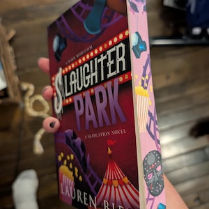 May include: A paperback book titled "Slaughter Park: A Slaycation Novel" by Lauren Biel. The cover features a carnival scene with a red and white striped tent, popcorn, and a purple roller coaster. The spine is pink with illustrations.
