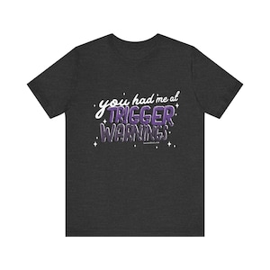 May include: Dark heather grey t-shirt with a purple graphic that says "You had me at trigger warnings".