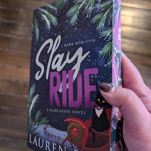 May include: A book cover with a dark blue background and a purple title that reads "Slay Ride" with the subtitle "A Slaycation Novel." The cover features a black cat sitting in a red sleigh with a skull and crossbones on the side. The book is by Lauren Biel.