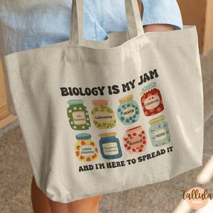 May include: Beige canvas tote bag with the text "BIOLOGY IS MY JAM" and colorful illustrations of jam jars. Each jar has a different label, including "Life Sciences," "Mentor," and "Passion." The phrase "AND I'M HERE TO SPREAD IT" is also printed.
