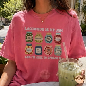 May include: A pink short-sleeve t-shirt featuring the text LACTATION IS MY JAM AND I'M HERE TO SPREAD IT. The graphic displays eight colorful jars labeled with terms including Milk Supply, Collaboration, Breast Pump, and Donor Milk.