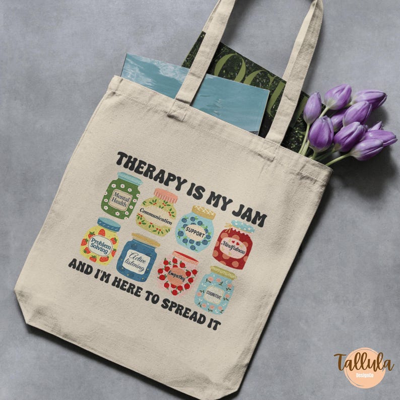 Puede incluir: Bolso tote beige con el texto "THERAPY IS MY JAM" y "AND I'M HERE TO SPREAD IT". La bolsa presenta ilustraciones coloridas de tarros de mermelada etiquetados con conceptos de salud mental. Se ve un ramo de tulipanes morados.