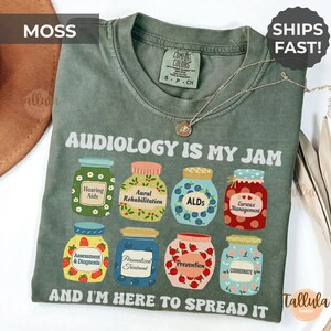 May include: Moss green t-shirt with the text "AUDIOLOGY IS MY JAM" and illustrations of jam jars. Each jar represents an audiology concept, such as "Hearing Aids" and "Earwax Management". The shirt also says "AND I'M HERE TO SPREAD IT".