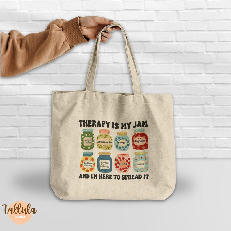 Puede incluir: Un bolso tote beige con el texto "THERAPY IS MY JAM" y "AND I'M HERE TO SPREAD IT". El dise&ntilde;o presenta frascos coloridos con etiquetas como "Mental Health" y "Communication". El bolso lo sostiene una persona.