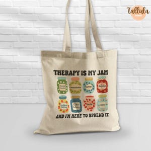 Puede incluir: Un bolso tote beige con el texto "THERAPY IS MY JAM" y "AND I'M HERE TO SPREAD IT". El dise&ntilde;o presenta botes de mermelada coloridos con etiquetas como "Comunicaci&oacute;n" y "Apoyo".