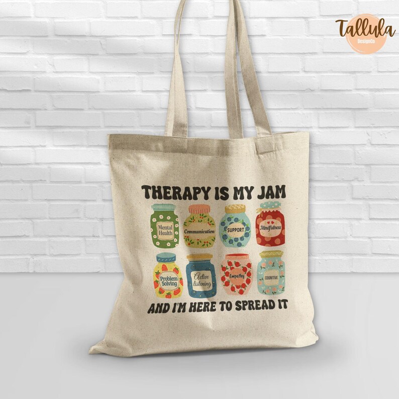Puede incluir: Un bolso tote beige con el texto "THERAPY IS MY JAM" y "AND I'M HERE TO SPREAD IT". El dise&ntilde;o presenta botes de mermelada coloridos con etiquetas como "Comunicaci&oacute;n" y "Apoyo".
