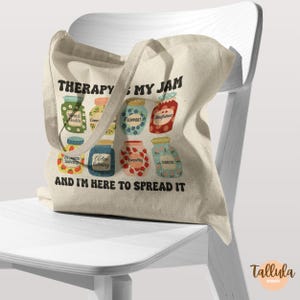 Puede incluir: Una bolsa de lona beige con el texto "THERAPY IS MY JAM AND I'M HERE TO SPREAD IT." La bolsa presenta ilustraciones coloridas de tarros de mermelada con etiquetas como "Mental Health" y "Support".