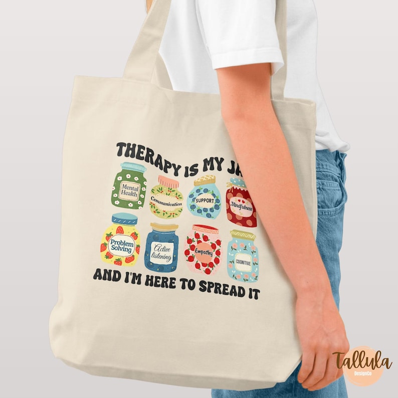 Puede incluir: Bolso tote color crema con el texto "THERAPY IS MY JAM" y "AND I'M HERE TO SPREAD IT." El bolso presenta ilustraciones de tarros de mermelada etiquetados con t&eacute;rminos de salud mental como "Support" y "Empathy."