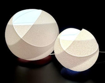 Op de afbeelding: Twee witte bolvormige lampen met een gestructureerd oppervlak. De grotere lamp is op de achtergrond en de kleinere lamp is op de voorgrond. Beide lampen zijn verlicht.