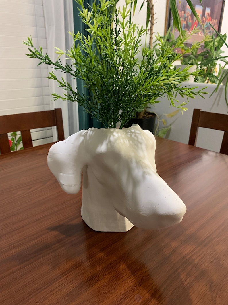 May include: White ceramic dog head vase with green leafy plant inside. The vase is sitting on a wooden table.