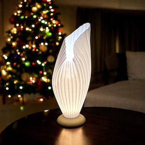 Può includere: Lampada da tavolo in ceramica bianca con un design unico e pieghettato. La lampada è accesa e proietta una luce calda.