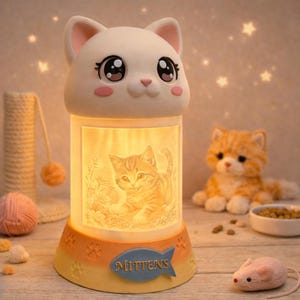 May include: A white cat-shaped night light with large eyes and pink cheeks. The cylindrical body displays a kitten image. The base is orange and yellow with paw prints and a blue fish with the word "Mittens".