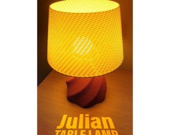 May include: A yellow table lamp with a woven shade and a red geometric base. The lamp is turned on and the light is shining. The text "Julian TABLE LAMP" is visible on the surface below the lamp.