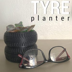 Pode incluir: Um vaso de pneu preto com uma planta suculenta crescendo nele. O vaso é feito de dois pneus empilhados. Um par de óculos de armação preta com hastes vermelhas está sobre uma mesa na frente do vaso. O texto "TYRE planter" está escrito em letras brancas sobre um fundo branco.