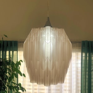 May include: A white, geometric pendant light fixture with a pleated paper shade. The light is hanging from a white cord and is illuminated, casting a warm glow.