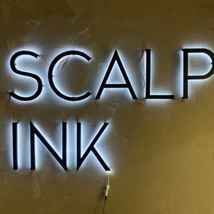 May include: A white neon sign with black lettering that reads "SCALP INK" mounted on a beige wall.