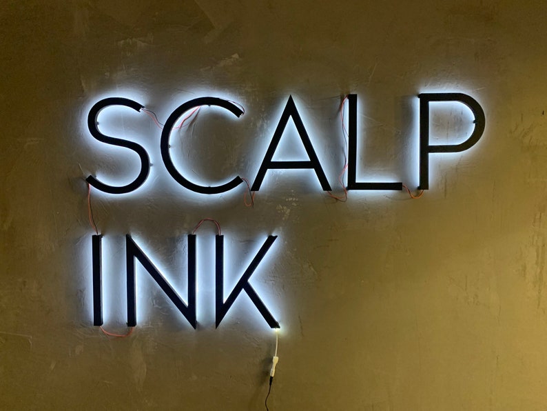 May include: A white neon sign with black lettering that reads "SCALP INK" mounted on a beige wall.