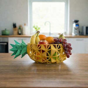 May include: A yellow pineapple-shaped fruit bowl filled with fresh fruit, including bananas, oranges, grapes, and an apple. The bowl has a geometric design and is placed on a wooden table. The green pineapple top adds a decorative touch.