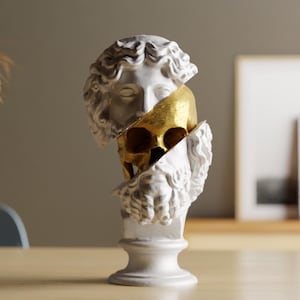 Puede incluir: Una escultura de busto blanco con un interior de calavera dorada. La escultura es de una figura clásica, dividida para revelar la calavera. El busto está sobre un pedestal blanco, sobre una mesa de madera.