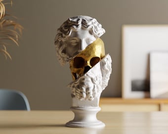 Op de afbeelding: Een witte borstbeeldsculptuur met een gouden schedelinterieur. De sculptuur is van een klassieke figuur, gesplitst om de schedel te onthullen. Het borstbeeld staat op een witte sokkel, op een houten tafel.