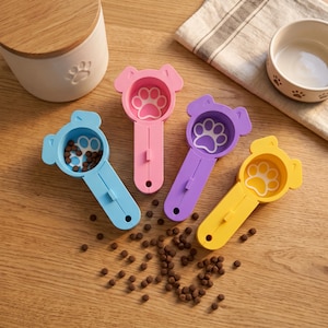 May include: Four plastic pet food scoops in blue, pink, purple, and yellow, each shaped like a dog head with a paw print design in the bowl. The blue scoop contains small brown kibble pieces, and loose kibble is scattered on the surface below.