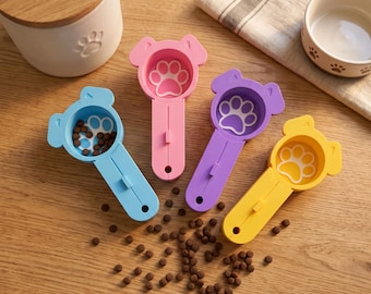 May include: Four plastic pet food scoops in blue, pink, purple, and yellow, each shaped like a dog head with a paw print design in the bowl. The blue scoop contains small brown kibble pieces, and loose kibble is scattered on the surface below.