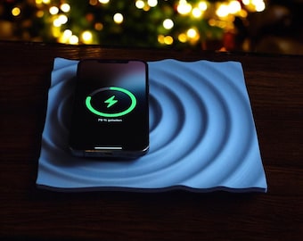 May include: A black smartphone charging wirelessly on a light blue, wave-patterned charging pad. The phone screen displays a green charging symbol and the text "79% charged". The background features blurred, warm-toned lights.