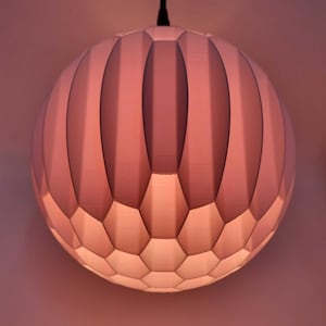 Puede incluir: Una lámpara colgante esférica rosa con un diseño geométrico. La luz tiene una serie de crestas verticales y redondeadas en la mitad superior y un patrón de panal en la mitad inferior. Está suspendida de un cable negro.