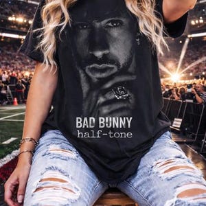 May include: Black t-shirt featuring a grayscale image of Bad Bunny with the text "BAD BUNNY half-tone". The shirt is worn with light blue ripped jeans. The background shows a stadium setting.