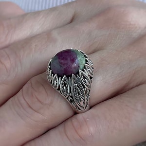 Op de afbeelding: Een zilveren ring met een grote, ronde, groene en roze edelsteen gezet in een gedetailleerd, bladerig ontwerp.