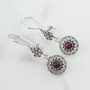 May include: A pair of silver earrings with a filigree design and a round, dark purple gemstone in the center of each earring.