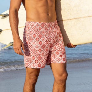 Puede incluir: Pantalones cortos de baño con estampado rojo y blanco. Los pantalones cortos presentan un diseño geométrico repetitivo. El modelo está de pie en una playa con una tabla de surf.