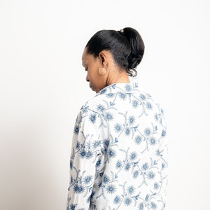 Può includere: Camicia bianca con motivo floreale blu. La camicia a maniche lunghe presenta un delicato motivo di fiori e steli blu. La camicia è abbinata a jeans blu scuro. La modella è di spalle.