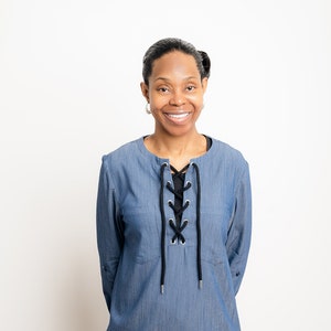 Può includere: Una camicia blu in stile denim con maniche lunghe e scollatura allacciata. La camicia ha un orlo arrotondato ed è abbinata a pantaloni neri. Il dettaglio allacciato nero aggiunge un elegante contrasto al tessuto blu.