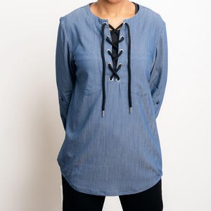 Può includere: Una camicia di jeans blu con maniche lunghe e scollatura allacciata. La camicia ha un colletto rotondo e un dettaglio di allacciatura nero. Le maniche sono arrotolate sui gomiti. La camicia è indossata con pantaloni neri.