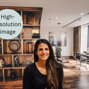 May include: A woman with long brown hair is standing in front of a bookshelf in a modern office. The bookshelf is made of wood and has several shelves filled with books and decorative items. The office has large windows, hardwood floors, and a desk with a chair.