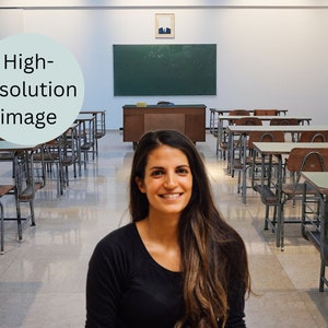 May include: A young woman with long brown hair smiles in a classroom setting. The room has rows of desks with chairs, a blackboard, and a teacher's desk.