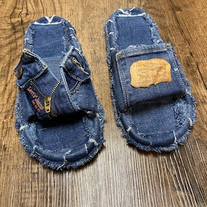 May include: A pair of blue denim slides with frayed edges. The slides have a zipper detail and a Levi's patch on the strap.