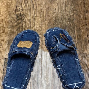 以下が含まれることがあります： フリンジの付いた青いデニムのスリッパのペア。片方のスリッパには、"Levi Strauss & Co."という文字が書かれた茶色の革のパッチが付いています。もう片方のスリッパにはジッパーのディテールがあります。