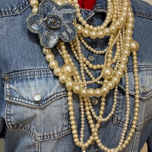 May include: A multi-strand necklace featuring cream-colored faux pearls of varying sizes layered over a blue denim shirt. A large, five-petaled flower brooch made from frayed blue denim fabric with a central beaded embellishment is attached.