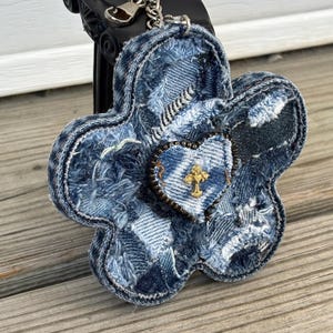 May include: A flower-shaped keychain crafted from distressed blue denim fabric patches with frayed edges. A small heart-shaped denim patch is centered on the flower, featuring a gold-toned cross charm and a dark beaded border, attached to a metal ring.