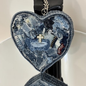 May include: A heart-shaped keychain crafted from various shades of blue denim fabric scraps with frayed edges. The surface features a small gold-toned cross charm embedded with clear rhinestones, attached to a silver-toned metal ring and chain.