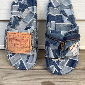 May include: A pair of slide sandals constructed from patchwork blue denim fabric with frayed edges. The left sandal features a tan Levi Strauss & Co. leather patch. The right sandal includes a metal zipper detail and a white 510 Skinny tag.