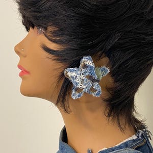 May include: A denim star-shaped earring is worn on a mannequin. The earring is made of blue denim fabric with white stitching and frayed edges. The mannequin has dark hair and is wearing a denim jacket.