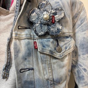 May include: A light blue acid-wash denim jacket layered over a grey hooded sweatshirt. A large, frayed denim flower brooch with a central white pearl and a small red Levi's brand tag is pinned to the chest pocket area of the denim jacket.
