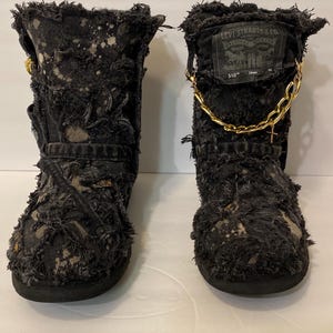 May include: A pair of black denim boots with a distressed look. The boots have a gold chain and a Levi's patch on the side. The boots are made of a soft, fuzzy material.