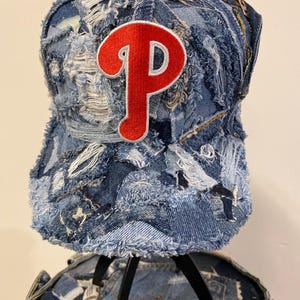May include: A distressed blue denim baseball cap featuring a patchwork design with frayed edges and exposed seams. A large, embroidered red letter P with a white outline is centered on the front panel of the cap, which is constructed from multiple shades of denim fabric.