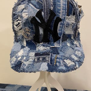 May include: A baseball cap constructed from multiple patches of blue denim fabric with frayed, distressed edges. The front panel features a prominent, embroidered black New York Yankees logo. The design incorporates various shades of blue denim.