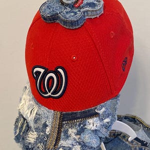 May include: A red baseball cap with a denim flower accent on top. The cap features the Washington Nationals logo in white and navy blue. The brim and back of the cap are embellished with distressed denim.