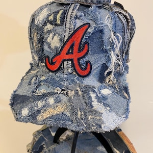 May include: A distressed denim baseball cap with a red "A" logo. The cap is made from patchwork denim with frayed edges, giving it a unique, worn look. The hat is a classic six-panel design with a curved brim.